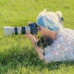 Woman with blue and white hair taking photos with a camera in a grassy field.